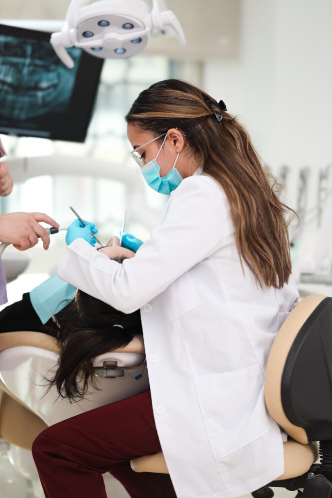 Dentiste femme portant une blouse blanche, un masque et des gants médicaux, réalisant un soin dentaire sur une patiente installée sur un fauteuil dentaire, dans un cabinet moderne équipé d’un écran de radiographie et de matériel professionnel.