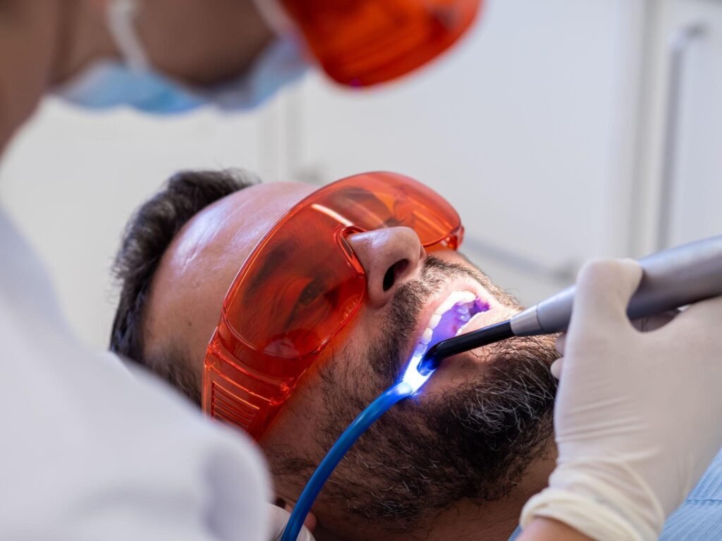 Séance de blanchiment dentaire en cabinet avec lampe LED activée sur les dents du patient, portant des lunettes de protection pour un traitement d’éclaircissement dentaire professionnel.