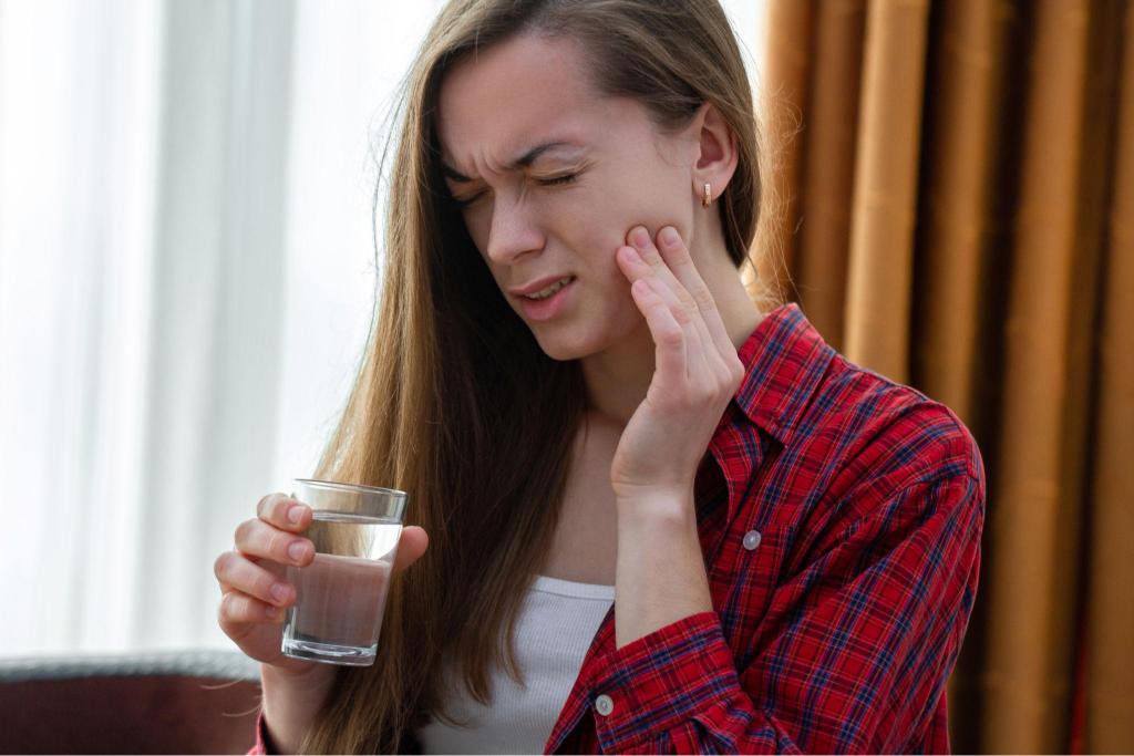 Jeune femme souffrant d'un mal de dent et tenant sa joue tout en essayant de boire un verre d'eau.